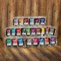 A set of packaged chocolate-covered snacks or bars arranged in three rows on a wooden surface against a wooden plank background. Each package has a different bright color label, and the chocolate pieces are visible through clear plastic bags.